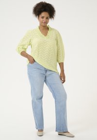 Suéter de color verde lima con un patrón en zigzag, cuello con volantes y mangas de tres cuartos, combinado con jeans de pierna recta en un tono azul claro.