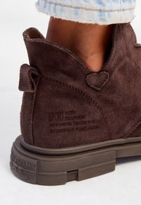 Ruske brune ankelstøvler i ruskind med hjerteformet detalje på hælen, iført med lyseblå denim jeans, på en hvid baggrund.