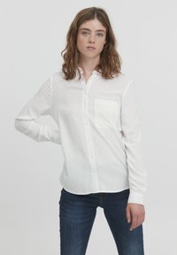 Hvid knap-up skjorte med en enkelt brystlomme, lange ærmer og et glat, let stof; båret med mørkeblå jeans.