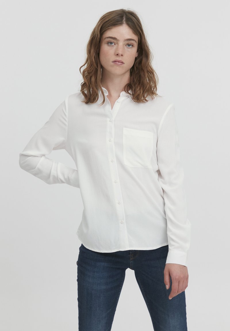 Hvid knap-up skjorte med en enkelt brystlomme, lange ærmer og et glat, let stof; båret med mørkeblå jeans.
