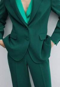 Femme portant un costume vert foncé ajusté avec un pantalon assorti et un chemisier vert plus clair en dessous, les mains dans les poches.