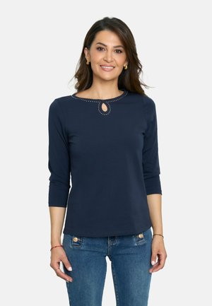 Camiseta de manga larga azul marino con cuello redondo que presenta un corte en forma de llave y tachuelas metálicas. Tejido suave, diseño ajustado y textura mínima.