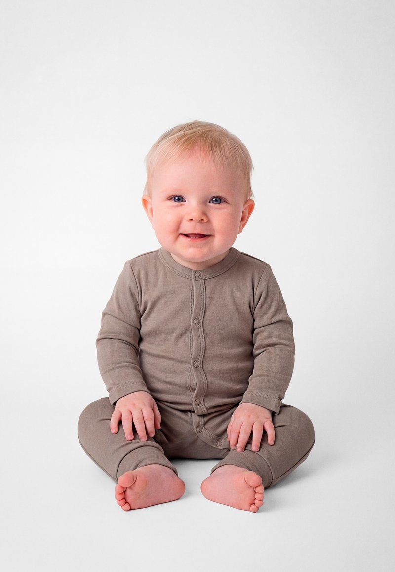 Bambino sorridente con occhi azzurri e capelli biondi seduto a gambe incrociate, indossa una tutina marrone con bottoni su sfondo bianco uniforme.