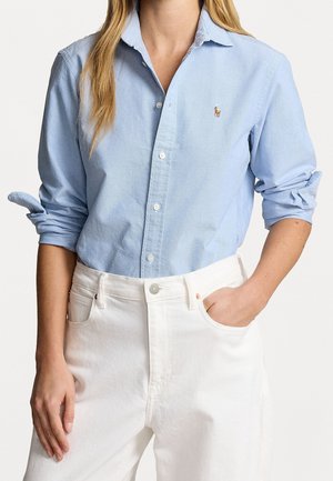 Femme portant une chemise bleu clair à boutons avec manches retroussées et un pantalon blanc taille haute, une main dans la poche, debout devant un fond uni.