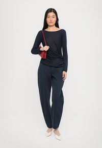 Marinblå långärmad topp med en vriden design, i kombination med marinblå vida byxor. Modellen håller en liten röd väska och bär spetsiga beige ballerinor.