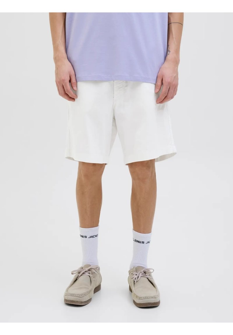 Pantalones cortos de algodón blancos con un ajuste cómodo, que presentan un corte recto y bolsillos laterales. Llevados con zapatos gris claro y calcetines blancos.