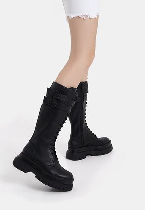 Bottes noires en cuir montant jusqu'au genou avec une semelle épaisse, un laçage à l'avant, des accents de double boucle et une fermeture éclaire latérale pour un enfilage facile.