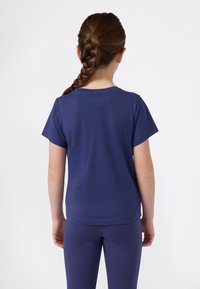 Niña con cabello trenzado castaño, vestida con una camiseta de manga corta azul marino y leggings a juego, de pie de espaldas contra un fondo blanco.