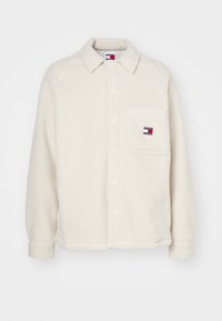 Chaqueta de camisa de felpa color crema con cierre de botones, cuello puntiagudo, un bolsillo en el pecho y un pequeño parche con el logo en el bolsillo.