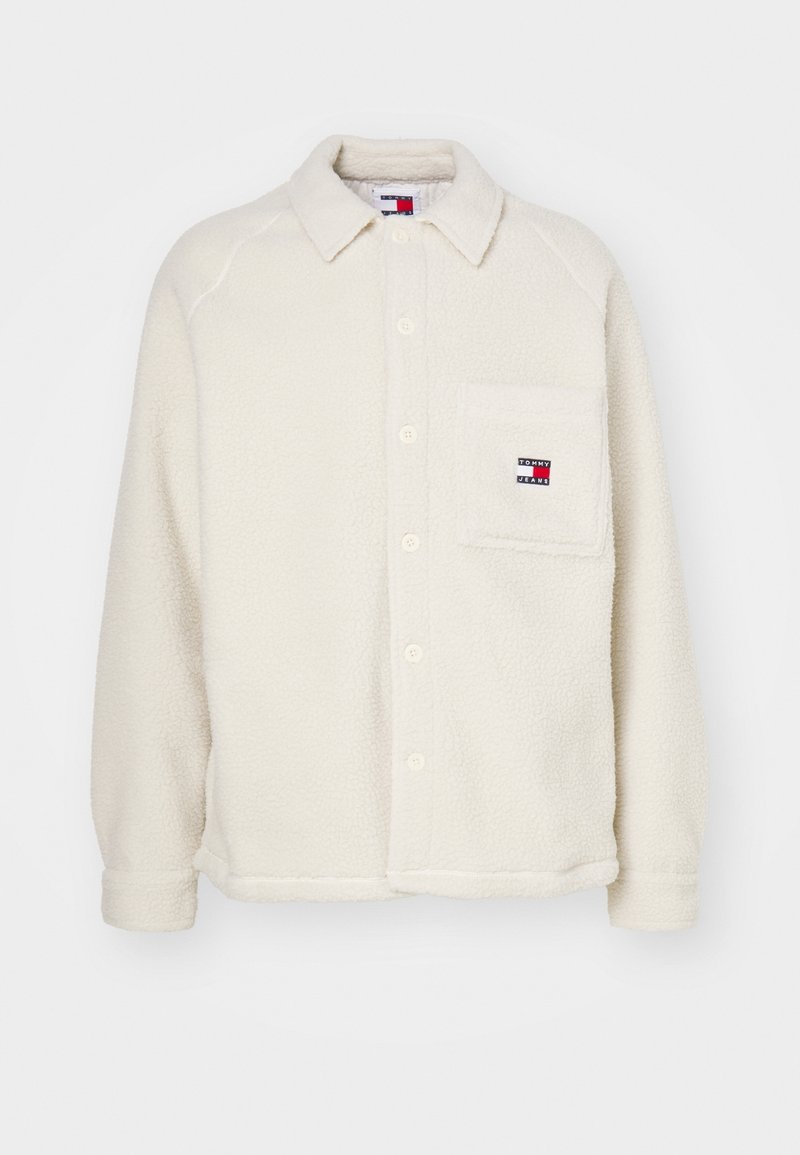 Chaqueta de camisa de felpa color crema con cierre de botones, cuello puntiagudo, un bolsillo en el pecho y un pequeño parche con el logo en el bolsillo.
