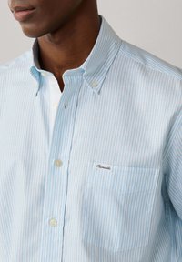 Homme portant une chemise à rayures bleu clair et blanches à boutons avec une poche poitrine étiquetée "Faconnable".