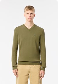 Jersey de punto verde con cuello en V, que presenta un diseño ajustado y puños y dobladillo acanalados. Logo de Lacoste bordado en el pecho.