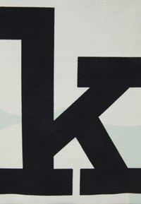 Lettre noire "K" sur un fond blanc texturé avec des accents bleu clair subtils. Le motif présente une texture tissée semblable à un nid d'abeilles.