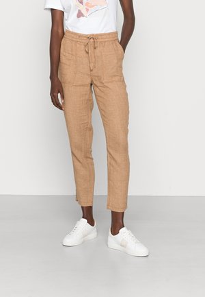 Person wearing tan drawstring linen pants with front pockets, cropped above ankles, paired with white sneakers and a white graphic t-shirt.