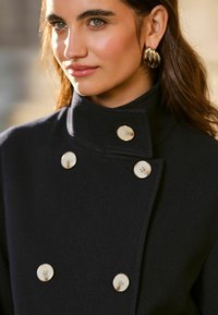 Cappotto di lana navy con colletto alto e design doppiopetto, accentuato da grandi bottoni crema. Texture liscia e silhouette su misura.