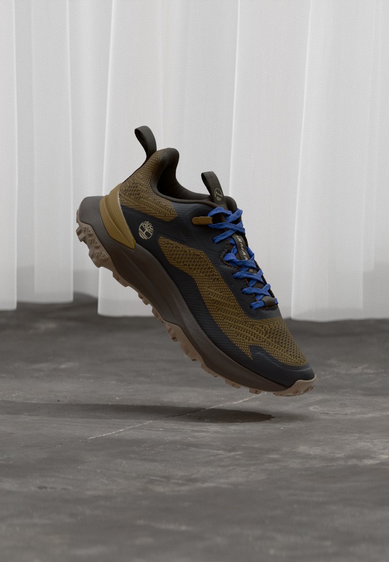 Athletischer Wanderschuh in Braun und Schwarz mit blauen Schnürsenkeln, erhoben über einem grauen Betonboden, weiße transparente Vorhänge im Hintergrund.