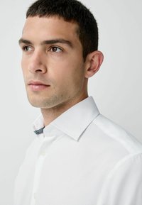 Valkoinen nappikauluspaida, jossa on jämäkkä kaulus ja hillitty tekstuurinen sininen yksityiskohta kauluksen reunassa. Sujuva kangas ja kapea muotoilu.