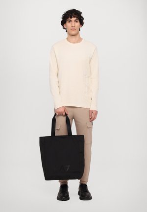 Borsa tote nera con logo in rilievo, manici in tessuto e forma rettangolare. Modello che indossa una maglietta a maniche lunghe color crema e pantaloni beige.