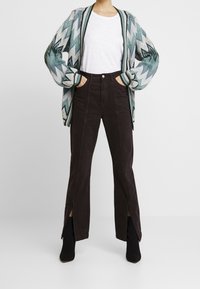 Person som bär en mönstrad blå-grå cardigan, vit t-shirt, högmidjade svarta byxor med framslutningar och svarta ankelboots, står med händerna i fickorna.