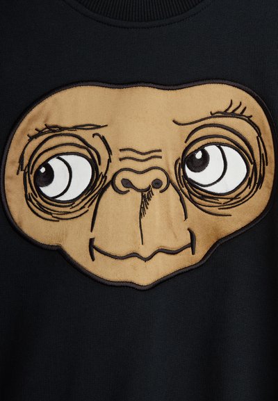 Μαύρη μπλούζα με κουκούλα που διαθέτει ένα καφέ κεντητό σχέδιο ενός καρτουνίστικου εξωγήινου προσώπου με μεγάλα μάτια και λεπτομερή χαρακτηριστικά προσώπου.