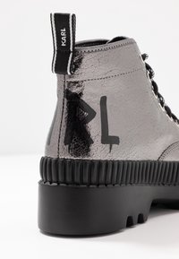 Botín de tobillo metálico negro y plateado con una textura agrietada, que presenta un prominente logo "KARL" y una suela de goma gruesa con nervaduras verticales.