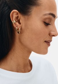 Gouden oorbellen in ringvormgetoond, waaronder een stevige manchetcuff en twee kleinere ringen, met een gepolijste afwerking en een gladde textuur, gedragen tegen een neutrale achtergrond.