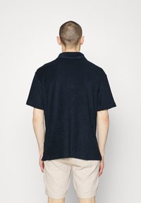 Man med kort hår klädd i en marinblå skjorta med kort ärm och beige shorts, stående vänd bort mot en enkel vit bakgrund.