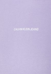Lavendelfärgad tyg med vitt "CALVIN KLEIN JEANS"-logotyp centrerad på det texturerade stickade materialet.