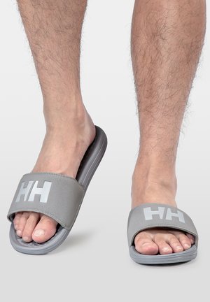 Piernas con sandalias deslizantes grises, cada sandalia presenta una correa ancha con "HH" impresa en blanco en la parte superior.