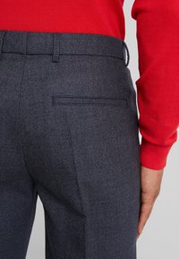 Gros plan d'une personne portant un pantalon gris foncé sur mesure et une chemise rouge à manches longues, un bras reposant le long du corps.