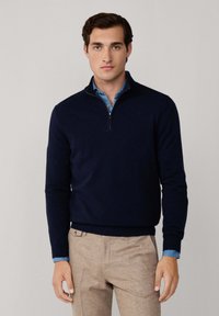 Junger Mann in einem marineblauen Pullover mit Viertel-Reißverschluss über einem blauen Hemd und beigen Hosen, stehend vor einem einfarbigen hellgrauen Hintergrund.