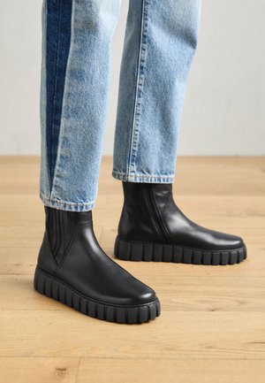 Personne portant un jean bleu clair et des bottines noires à semelles épaisses debout sur un sol en bois.