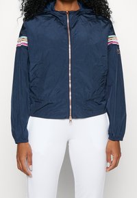 Námořnická modrá, lehká bunda s kapucí a plným zipem, se spoustou barevných pruhů na ramenech a elastickými manžetami.