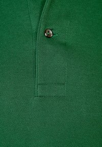 Žalias polo marškinėlis su tekstūruotu audiniu, apykakle ir vienu metaliniu mygtuku. Išskirtiniai siuvimo detalės ant apykaklės ir mygtukų juostos.