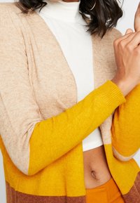 Vrouw met een beige, geel en bruin color-block cardigan over een witte cropped coltrui, waarbij haar buik zichtbaar is en haar opgeheven arm met een zichtbare oorring.