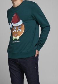 Sudadera verde oscuro con un gráfico de un hombre de jengibre que lleva un gorro de Santa y un corbata de moño. Corte clásico con puños y dobladillo de canalé.