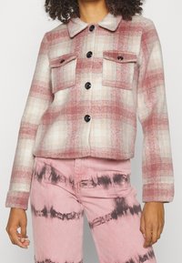 Ternet jakke i blødt, tekstureret stof med rosa og cremefarver. Har krave, to brystlommer og sorte knapper. Kombineret med pink tie-dye bukser.