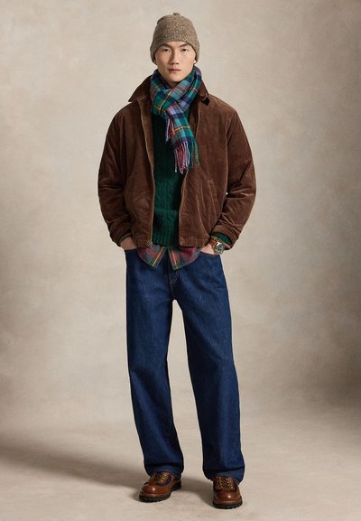 Mand iført brun kordjakke, grøn sweater, ternet skjorte, blå jeans, brune støvler, strikhue og blå-grøn ternet tørklæde, står med hænderne i lommerne.