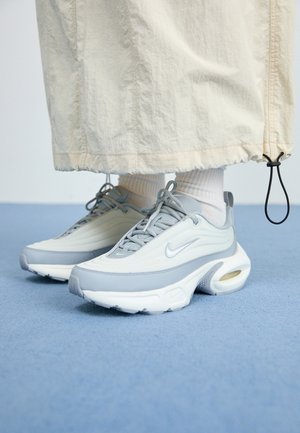 Feet wearing white socks and gray Nike Air Max sneakers with white and light beige wide pants on blue floor.