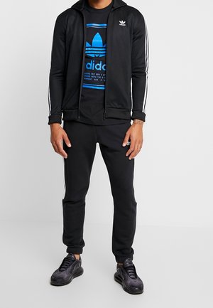 Survêtement Adidas noir comprenant une veste zippée avec des bandes blanches et un logo bleu, assortie à un pantalon de jogging noir et des baskets foncées.