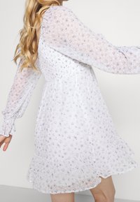 Vit blommig klänning med lila detaljer, genomskinliga långa ärmar, smockat liv och volangkant. Lättviktig tyg med delikat mönstring.