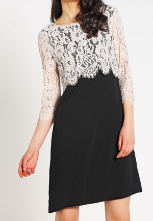 Femme portant une robe noire avec un haut en dentelle blanche à manches trois-quarts et bords festonnés.