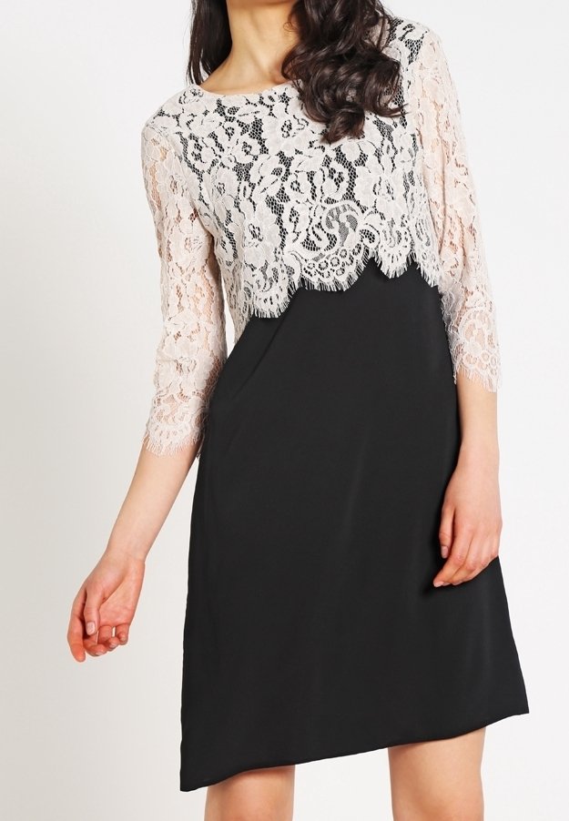 Femme portant une robe noire avec un haut en dentelle blanche à manches trois-quarts et bords festonnés.