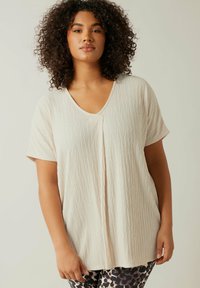 Lys beige kortærmet top med V-hals, der har et tekstureret, krøllet stof og en løs pasform. Kombineret med mønstrede sorte og beige leggings.