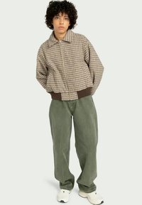 Bomber jacket a quadri marroni con chiusura a zip, polsini elasticizzati e orlo a coste marrone, abbinata a pantaloni in corduroy verde larghi e sneakers bianche.