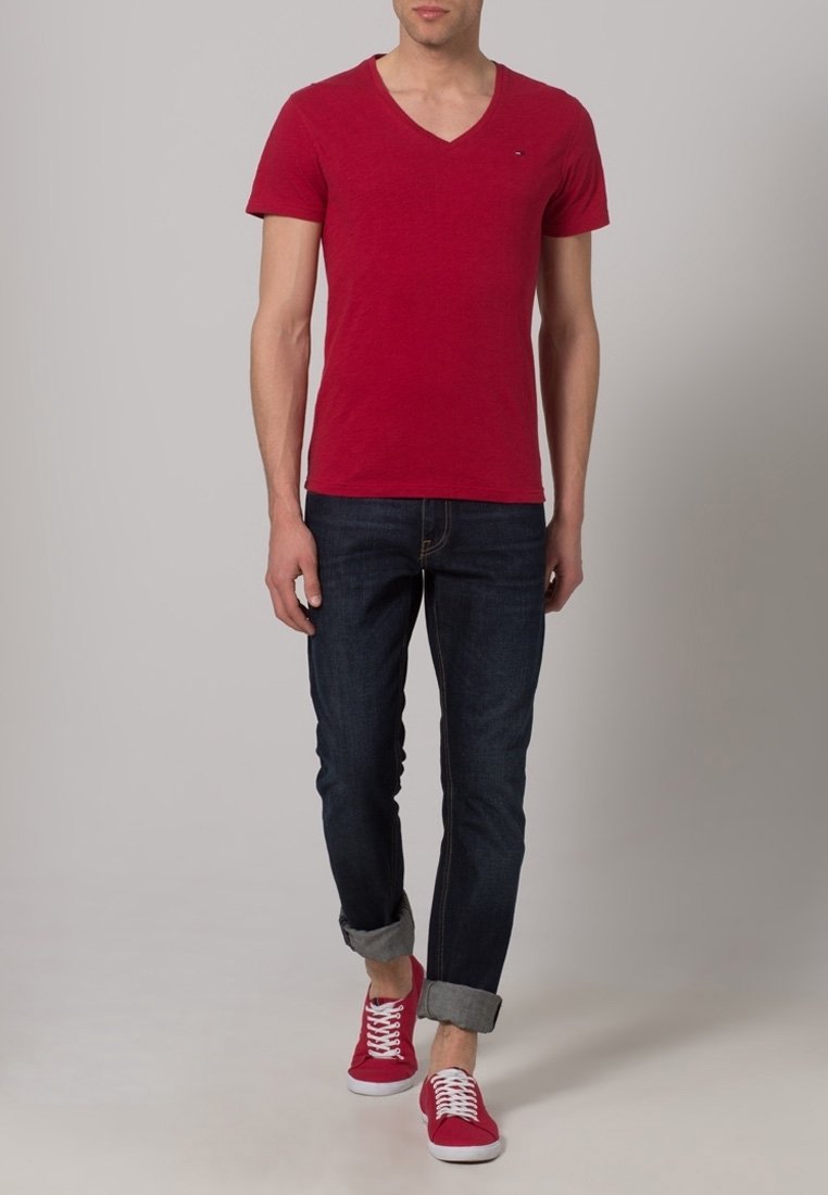 Homme portant un t-shirt rouge à col en V, un jean foncé retroussé et des baskets rouges, marchant devant un fond neutre.