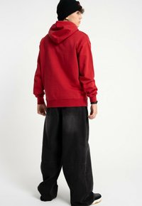 Czerwona bawełniana bluza z dużym kapturem, z prążkowanymi mankietami i dolnym końcem. W połączeniu z luźnymi czarnymi dżinsami z kieszeniami i swobodnym krojem. Czarna czapka beanie.