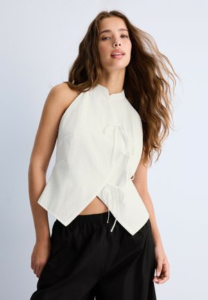 Jeune femme aux longs cheveux bruns portant un top blanc texturé sans manches avec des détails de lien et un pantalon noir ample, sur un fond uni.