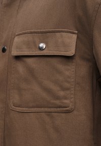 Barna szövetkabát zsebének fedele, amely egy kerek ezüstszínű fémgombbal van rögzítve, amelyen a "Jack & Jones" felirat látható.