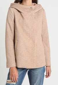 Beige, tekstureret uldfrakke med en stor hætte, der har to sidelommer og trykknaplukning. Bæres over slidte blå jeans.